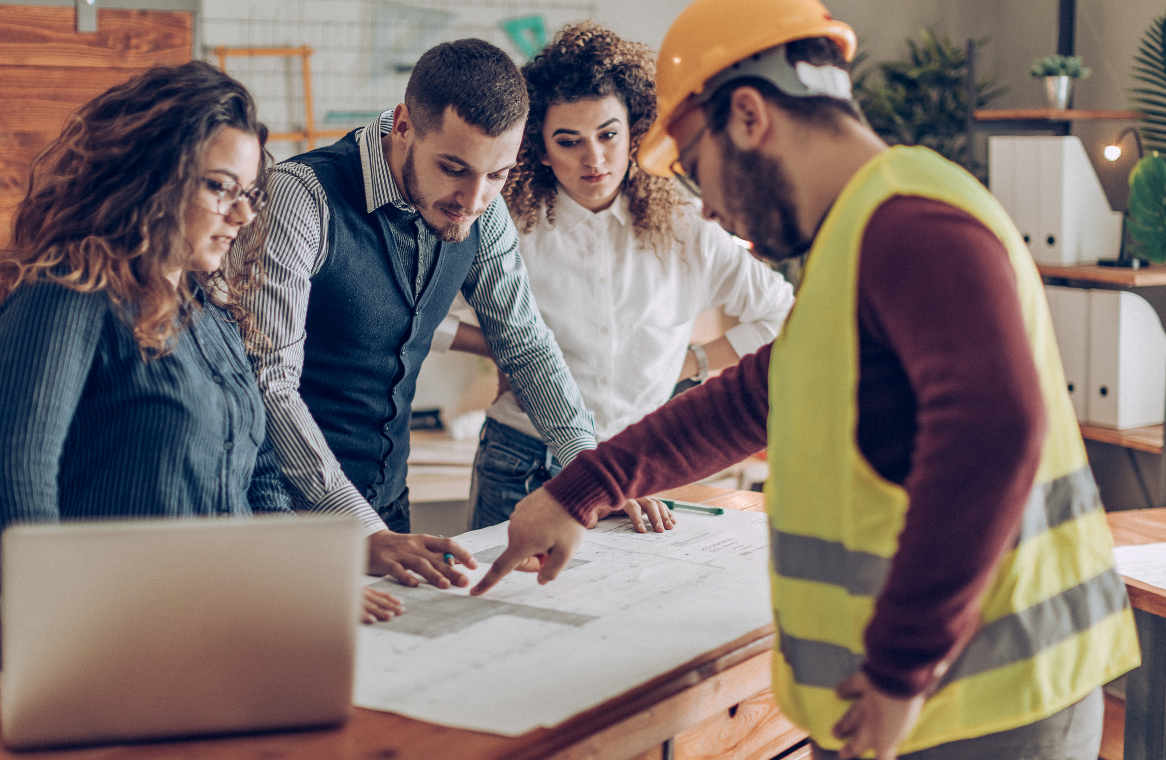 A group of professionals and a construction worker in a safety vest reviewing architectural blueprints on a wooden table.