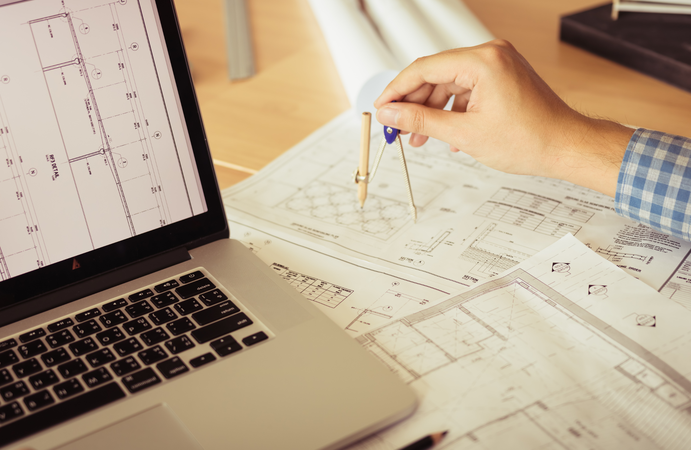 Architect reviewing building blueprints on desk with laptop displaying CAD drawings and drafting compass