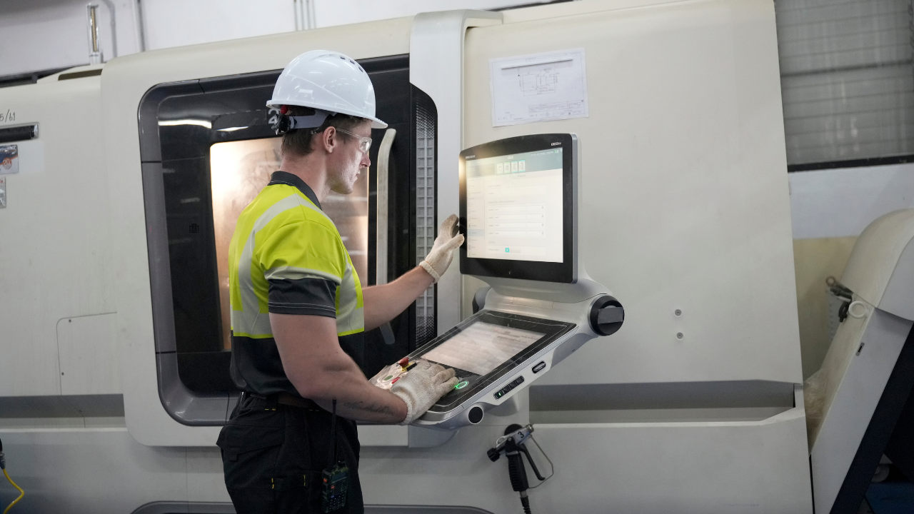 A manufacturing technician in a white hard hat and high-visibility vest operating a CNC machine control panel in an industrial facility
