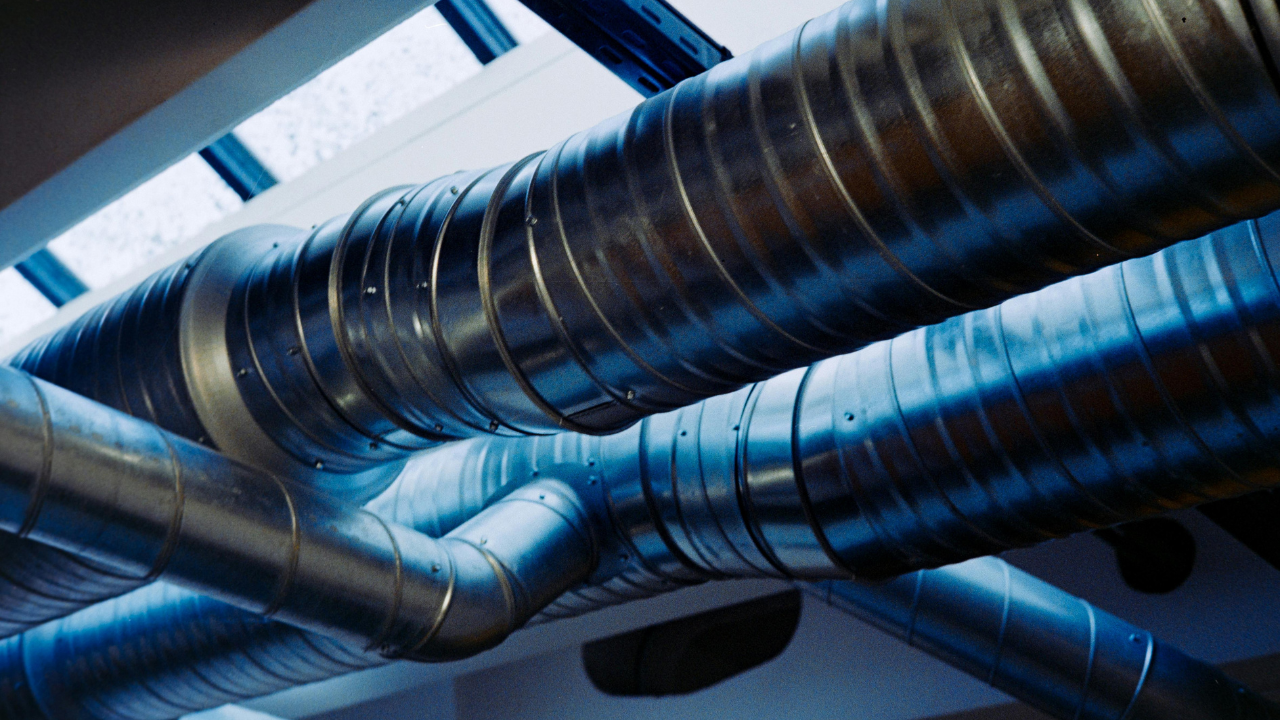 Close-up view of industrial HVAC ductwork with reflective metal pipes running across a ceiling beneath skylights.
