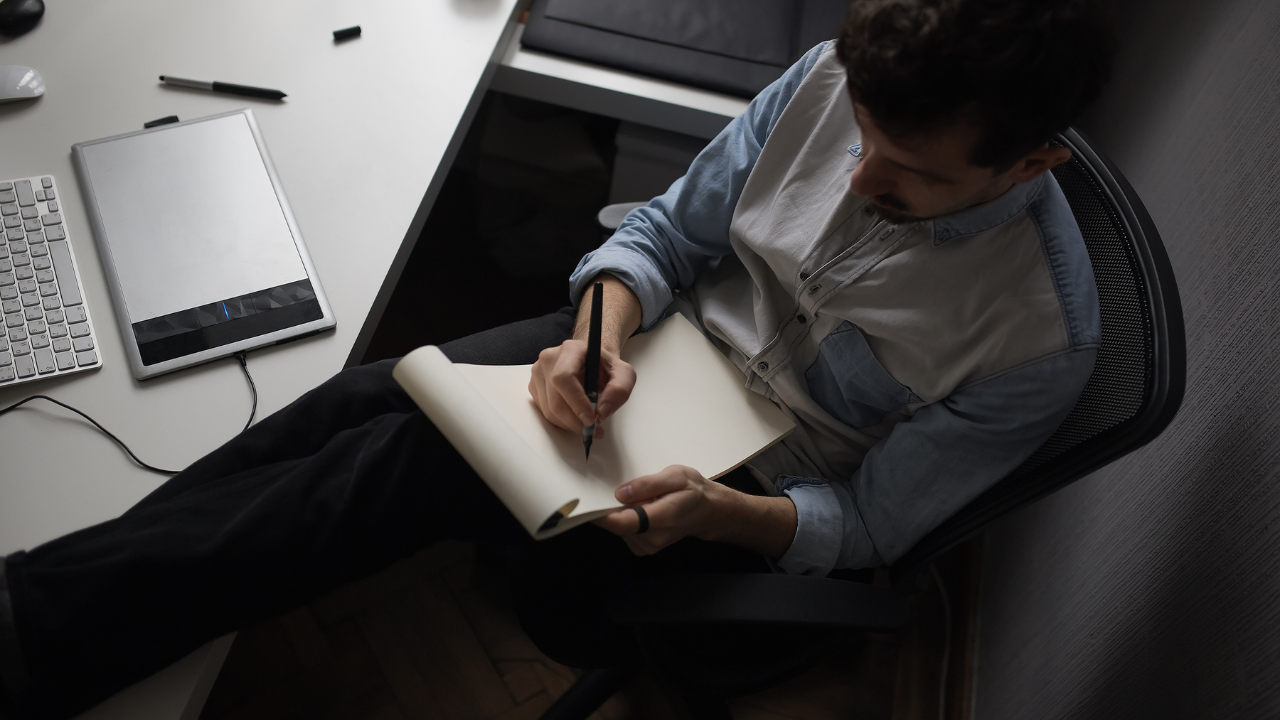 Man sitting at a desk with a tablet and keyboard nearby, writing notes in a blank notebook.